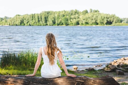back view of girl sitting on trunk of tree on river shore and looking awayの写真素材