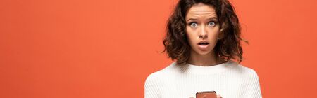 panoramic shot of shocked woman in autumnal outfit using smartphone isolated on orangeの写真素材