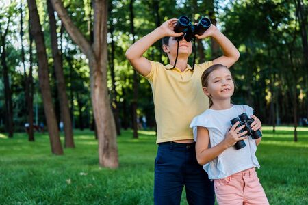 happy boy looking through binoculars near cute friend in parkの写真素材