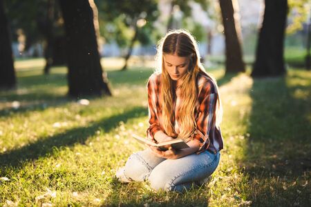 full length view of beautiful girl in casual clothes sitting on meadow in sunlight and reading bookの写真素材