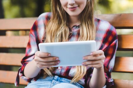 cropped view of young girl in casual clothes sitting on wooden bench in park and using digital tabletの写真素材