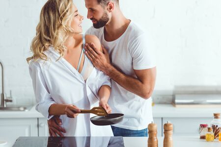 handsome young man hugging cheerful girlfriend preparing breakfast on frying panの写真素材