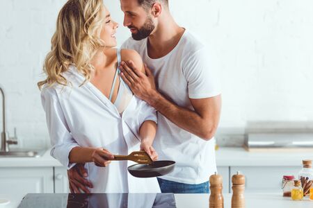 handsome young man hugging cheerful girlfriend preparing breakfast on frying pan