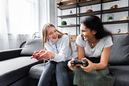 KYIV, UKRAINE - JULY 10, 2019: multicultural friends sitting on sofa and playing video game in...
