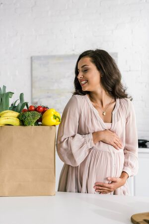 happy pregnant woman looking at paper bag with groceries while touching bellyの写真素材