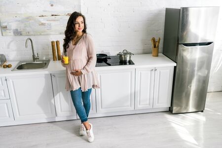 cheerful pregnant woman standing and holding glass with fresh orange juice in kitchen