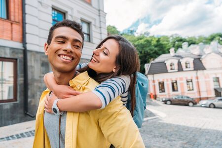 happy girl hugging cheerful mixed race man near buildings