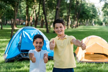 happy multicultural boys smiling while showing thumbs up near campsの写真素材