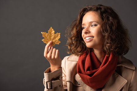 smiling woman in trench coat holding golden maple leaf on black backgroundの写真素材