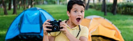 panoramic shot of surprised boy holding digital camera near camps in parkの写真素材
