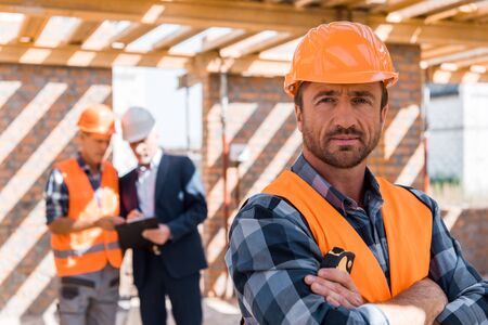 selective focus of handsome man in helmet standing with crossed arms near coworker and businessman