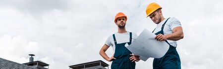 panoramic shot of architects standing with hands on hips near coworker with paperの写真素材