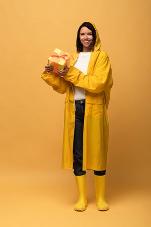 happy woman in yellow raincoat and wellies holding gift box on yellow backgroundの写真素材