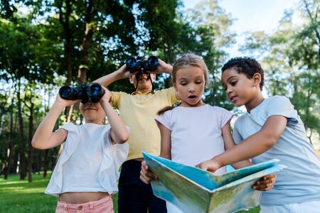 surprised kid looking at map with african american boy near friends with binoculars in park