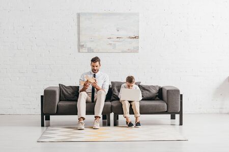 dad with toy plane and son with laptop sitting on sofaの写真素材