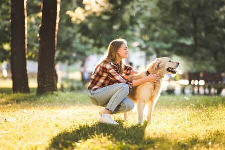 side view of beautiful girl in casual clothes petting golden retriever while sitting on meadow in...