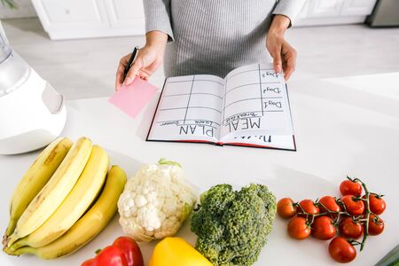 cropped view of woman near notebook with meal plan and vegetables on tableの写真素材