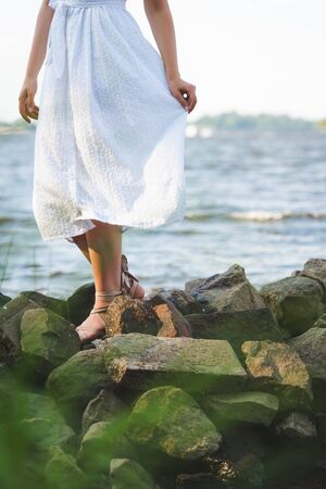 cropped view of girl in white dress walking on rocky river shoreの写真素材