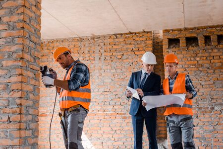 handsome builder using hammer drill near businessman and coworker