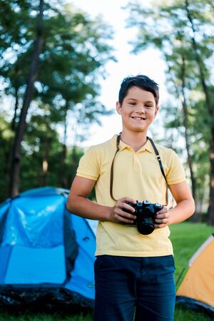 happy boy holding digital camera and smiling near camps in parkの写真素材