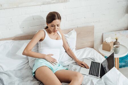 upset young woman lying on bed with laptop and touching bellyの写真素材