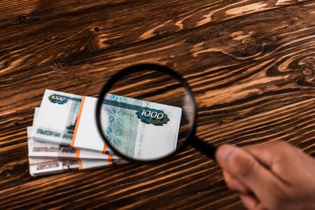 cropped view of man holding magnifier near russian money on wooden table
