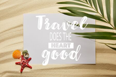 top view of green palm leaf near placard with travel does the heart good, starfish and seashells on sandy beachの写真素材