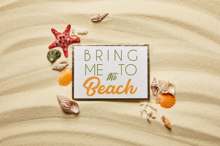top view of placard with bring me to the beach lettering near seashells, starfish and white corals on sandy beachの写真素材