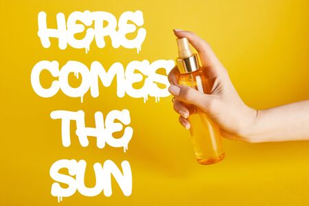 cropped view of woman holding bottle with sunscreen spray on yellow background with here comes the sun letteringの写真素材