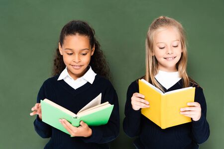 two smiling multicultural schoolgirls reading books while standing near green chalkboard