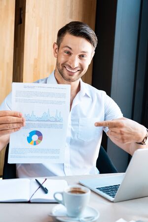 front view of smiling man pointing with finger at document with diagram in cafe