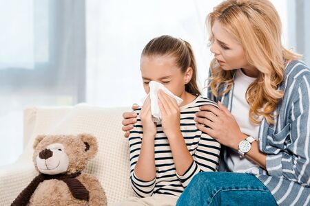 woman sitting near daughter sneezing in tissue near teddy bearの写真素材