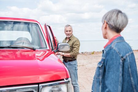 selective focus of senior couple of tourists standing near red car and looking at each other at beachの写真素材