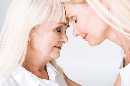 blonde mature daughter and senior mother standing face to face with closed eyes isolated on greyの写真素材