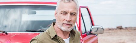 panoramic view of pensive senior man standing near car and looking away