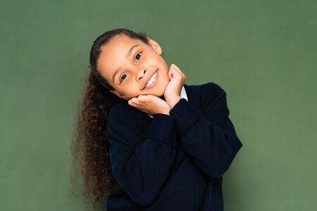 happy african american schoolgirl holding hands near face while standing near green chalkboard