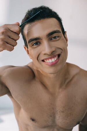 handsome mixed race man combing hair with hairbrush and smiling while looking at cameraの写真素材