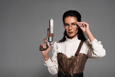 pensive steampunk woman holding vintage pistol and touching glasses isolated on greyの写真素材