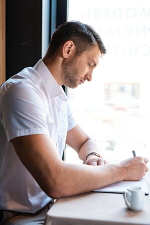 side view of focused handsome man writing in textbook in cafeの写真素材