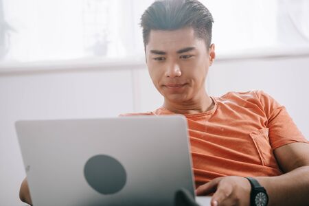 serious asian man using laptop while sitting on couch at home