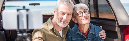 panoramic view of senior couple embracing and taking selfie near car
