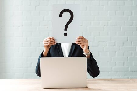 cropped view of woman covering face while holding board with question mark near brick wall and...