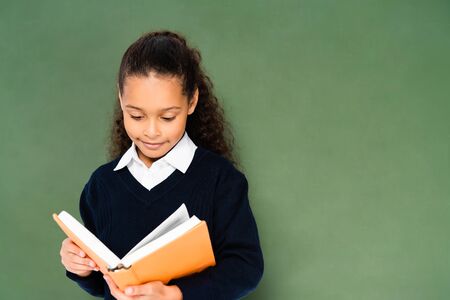 attentive african african american schoolgirl reading book while standing near chalkboardの写真素材