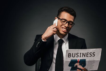 happy african american businessman in suit talking on smartphone while reading business newspaper on dark backgroundの写真素材