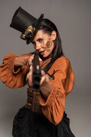 focused steampunk woman in top hat aiming with gun isolated on greyの写真素材