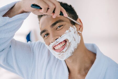 portrait shot of handsome man in bathrobe shaving face with razor blade, smiling and looking at cameraの写真素材