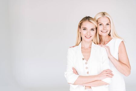 smiling blonde mature mother embracing young daughter in total white clothes isolated on greyの写真素材