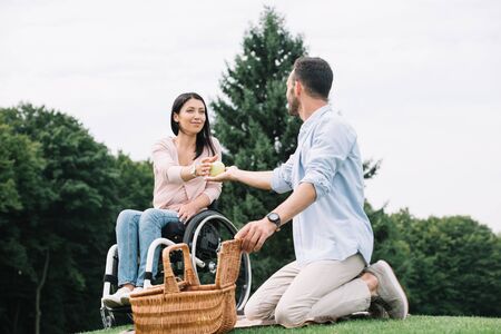young man giving apple to happy disabled girlfriend while resting together in parkの写真素材