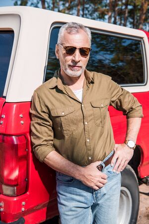 confident senior man in sunglasses standing with hand on hip near red car and looking at camera