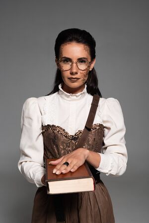 front view of steampunk woman in glasses holding book and looking at camera isolated on greyの写真素材